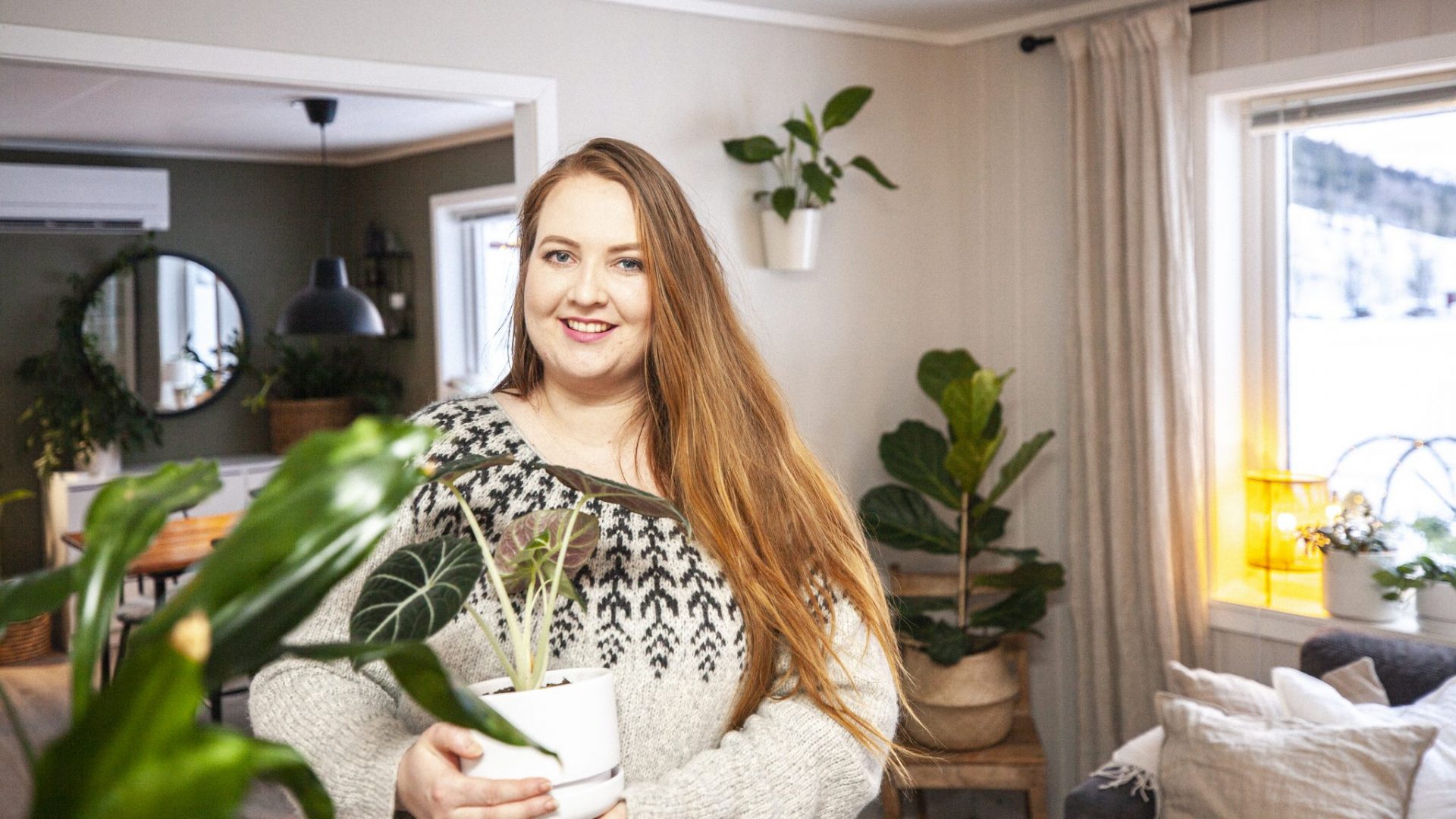 Synne står hjemme omgitt av grønne planter og holder en potteplante.