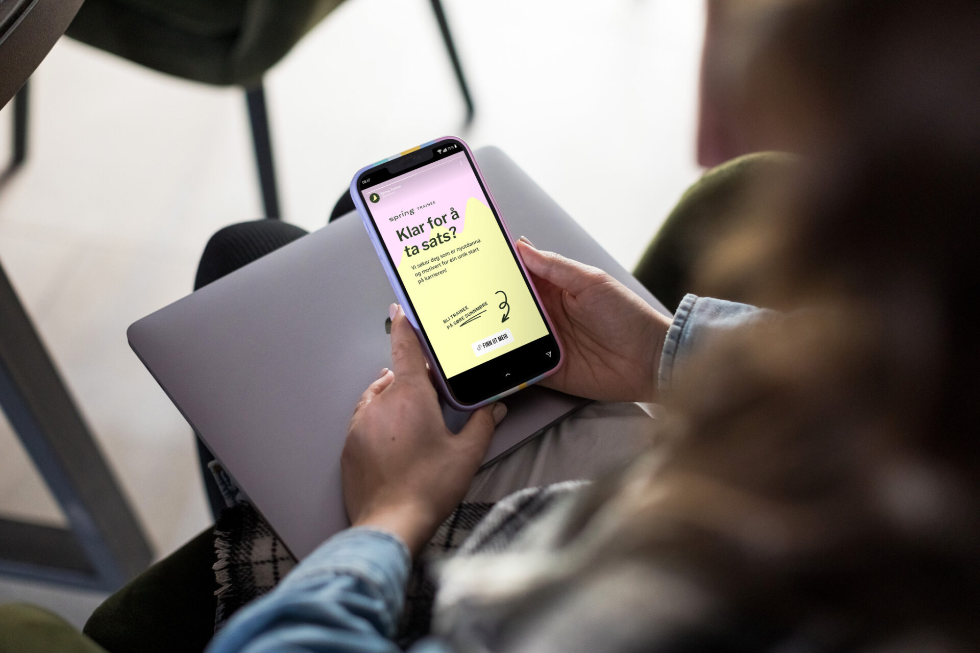 Person som holder en mobil med Spring Trainee-annonse med budskapet «Klar for å ta sats?».
