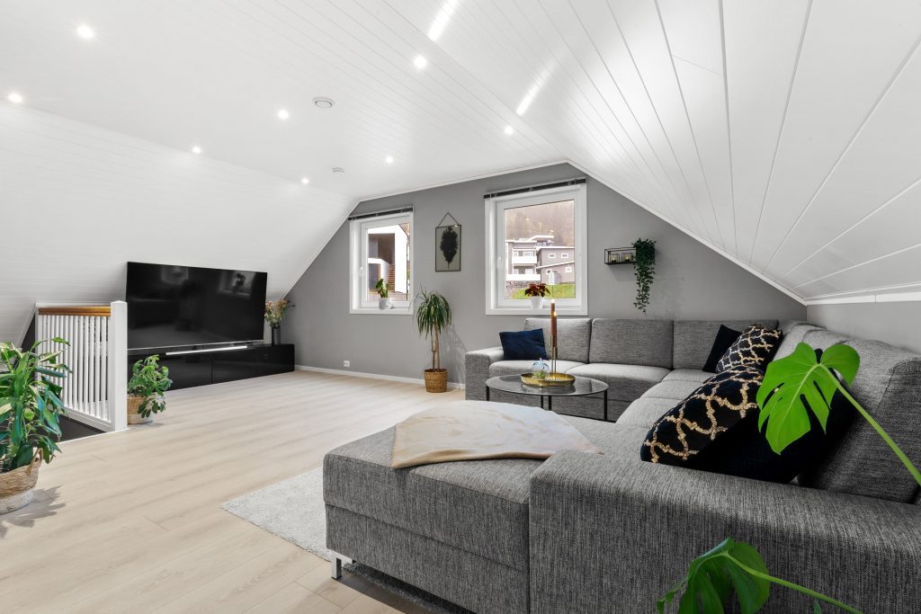 Lys loftstue med grå hjørnesofa, TV og grønne planter.